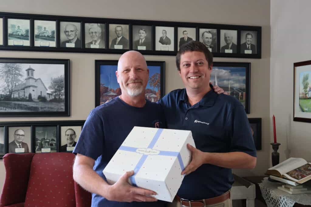 Two smiling men holding a gift box in a room with framed portraits and paintings on the walls at Fairview Church.