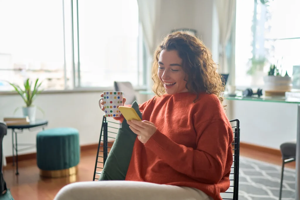 A woman in an orange sweater sits in a modern living room, holding a polka-dot mug and smiling at her smartphone, perhaps noticing some exciting changes on Google Search.